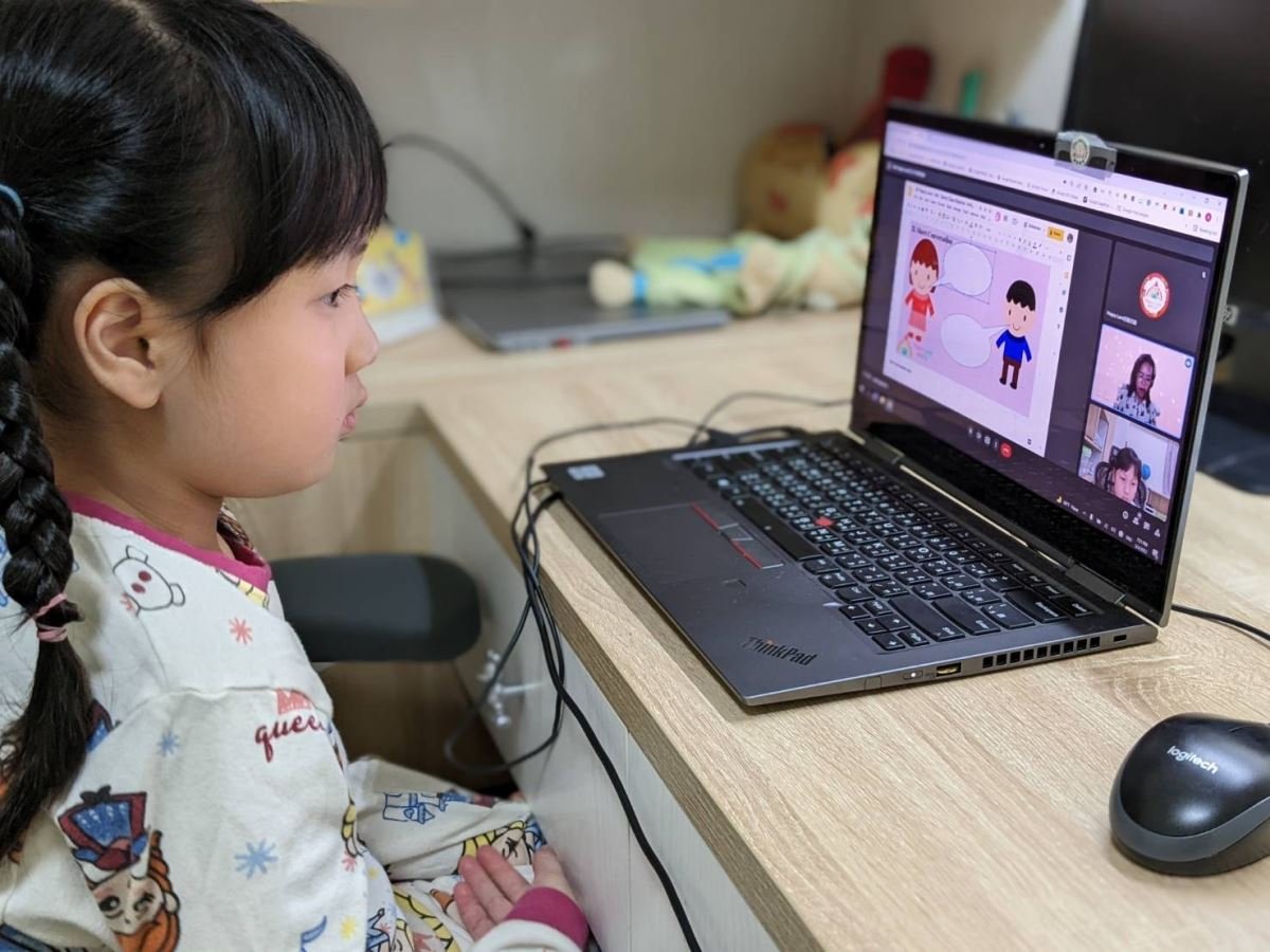 穿著睡衣的小女孩使用筆記型電腦參加線上英語課。 她專注於螢幕上顯示的卡通人物和其他孩子。