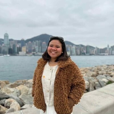 老師 Jasmin 在香港海濱微笑，背景是城市天際線。