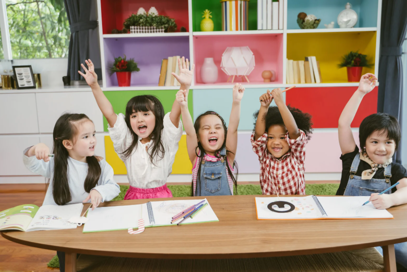 快樂的孩子們在色彩繽紛的課桌旁舉起手、學習和畫畫。 有效的早期英語學習。