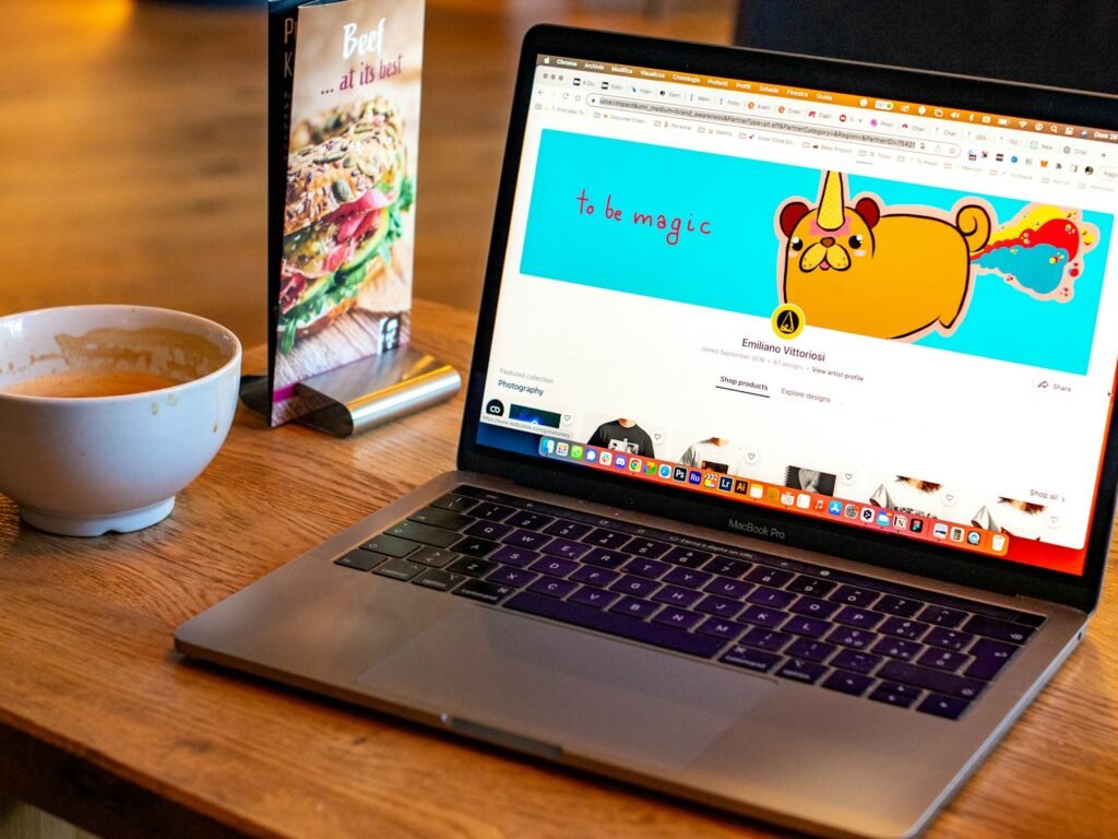 木桌上的MacBook Pro顯示線上設計作品，旁邊有咖啡和食物菜單。